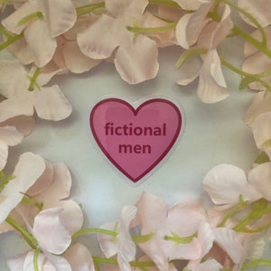 K&ouml;nnte beinhalten: Ein rosa herzf&ouml;rmiger Aufkleber mit den Worten "fictional men" in wei&szlig;em Text. Der Aufkleber ist von einem Kranz aus hellrosa Bl&uuml;tenbl&auml;ttern umgeben, was eine weiche, romantische &Auml;sthetik erzeugt.