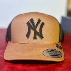 May include: A brown and black baseball cap with a black leather New York Yankees logo. The cap has a black mesh back and a brown brim.