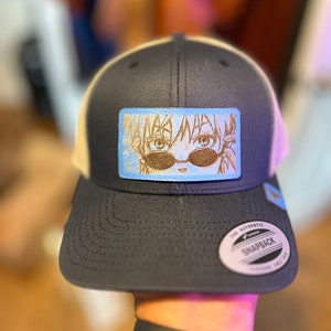 May include: A black and white trucker hat with a beige mesh back. The hat has a blue and white rectangular patch with a cartoon image of a person wearing sunglasses. The patch has the text "MAKE WAY" in a stylized font. The hat has a black "The Authentic Yupoong Snapback" label on the back.