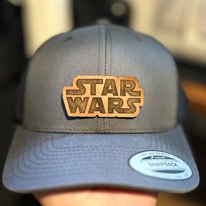 May include: A gray and black trucker hat with a brown wooden patch that says "STAR WARS" in a stylized font.
