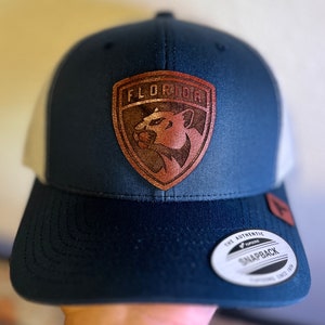 Può includere: Un cappello da camionista blu e bianco con una toppa in pelle marrone che presenta la parola "FLORIDA" e una pantera. Il cappello ha un'etichetta nera "Yupoong Snapback" sul retro.