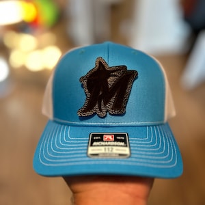 May include: A blue and white trucker hat with a black leather patch featuring a stylised letter "M". The hat has a white mesh back and a black label with the text "EST. 1970 RICHARDSON 112".
