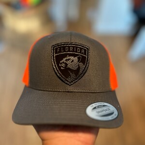 May include: A gray and orange baseball cap with a black leather patch that says "Florida" and features a panther. The patch is stitched onto the front of the cap. The cap has a black mesh back and a white "Snapback" label.