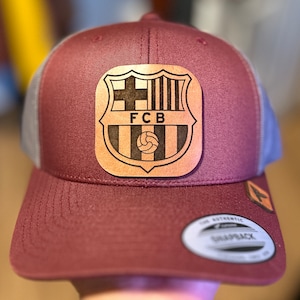 May include: A maroon and gray trucker hat with a brown leather patch featuring the FC Barcelona logo. The patch has a black and white design with the team's initials, "FCB", in the center.
