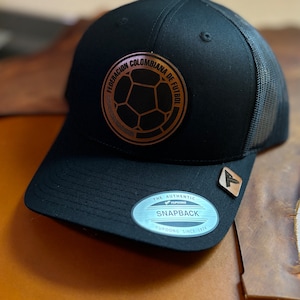 Puede incluir: Gorra de béisbol negra y marrón con un parche bordado en oro que dice "Federacion Colombiana de Futbol" y un balón de fútbol. La gorra tiene una parte trasera de malla y un cierre de presión negro. La gorra también tiene una pegatina plateada que dice "The Authentic Yupoong Snapback Yupoong Since 1974."