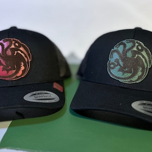 Pode incluir: Dois bonés de caminhoneiro pretos com um logotipo de dragão de três cabeças. O logotipo do boné da esquerda é rosa e vermelho, enquanto o logotipo do boné da direita é verde e azul.