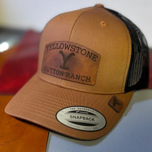 May include: A brown and black trucker hat with a leather patch that says "Yellowstone Dutton Ranch". The hat has a black mesh back and a brown front with a white logo that says "The Authentic Yupoong Snapback Yupoong Since 1974".