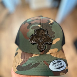 May include: A camouflage green, black, and brown baseball cap with a black mesh back. The cap has a brown leather patch with a tiger design on the front. The cap has a white label on the side that says "The Authentic Yupoong Snapback Since 1974".