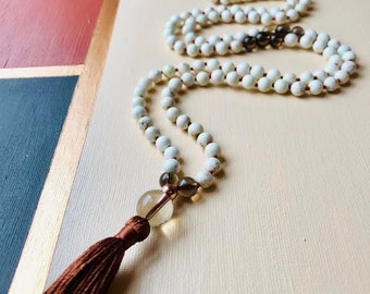 May include: White Howlite mala with a brown tassel and a clear quartz crystal pendant. The mala is made of small, round beads and is strung on a red thread.