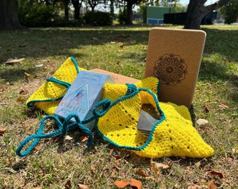 May include: A bright yellow crocheted pouch with teal trim and long ties, containing a deck of cards. Next to it sits a light blue box labelled The Starseed Oracle by Rebecca Campbell and Danielle Noel, and a rectangular cork yoga block with a mandala.