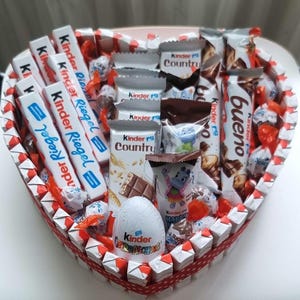 Può includere: Un cesto regalo a forma di cuore pieno di vari prodotti al cioccolato Kinder. Il cesto include barrette Kinder Riegel, barrette Kinder Bueno, barrette Kinder Country e un uovo Kinder Sorpresa. Il cesto è decorato con accenti rossi e bianchi.