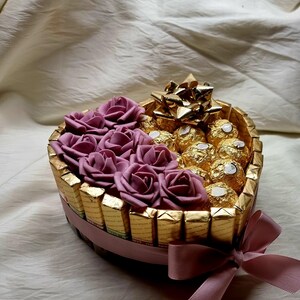May include: A heart-shaped box filled with chocolate candies and decorated with pink fabric roses. The box is tied with a pink ribbon.
