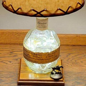 May include: A unique lamp made from a repurposed Blanton's bourbon bottle. The clear glass bottle is illuminated with lights and sits on a wooden base. A bronze horse and rider figurine is on the base. The lampshade is tan with a brown stitched trim.