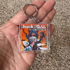 Pode incluir: Um chaveiro de acr&iacute;lico transparente com a arte da capa de "hack//G.U. Vol.1 Rebirth". A arte mostra dois personagens de anime em um fundo laranja e vermelho. O chaveiro tem um anel de metal prateado.