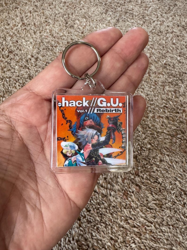 Pode incluir: Um chaveiro de acr&iacute;lico transparente com a arte da capa de "hack//G.U. Vol.1 Rebirth". A arte mostra dois personagens de anime em um fundo laranja e vermelho. O chaveiro tem um anel de metal prateado.