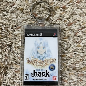 May include: Clear acrylic keychain featuring a PlayStation 2 game cover for .hack//INFECTION Part 1. The artwork shows a female character with long white hair and a group of smaller figures. The keychain has a silver metal ring.