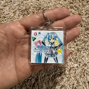 May include: A square clear acrylic keychain featuring a printed graphic of the character Hatsune Miku with long teal hair. The design includes the text Hatsune Miku Project DIVA f, along with red, blue, and green geometric gaming symbols.