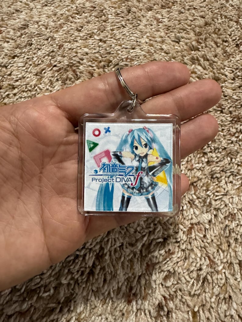 May include: A square clear acrylic keychain featuring a printed graphic of the character Hatsune Miku with long teal hair. The design includes the text Hatsune Miku Project DIVA f, along with red, blue, and green geometric gaming symbols.
