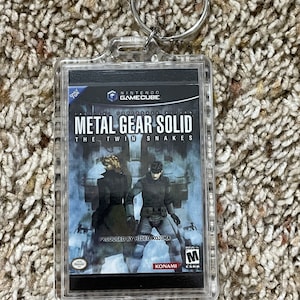 May include: Clear acrylic keychain featuring the Metal Gear Solid: The Twin Snakes video game cover art. The keychain includes a silver metal ring and the Nintendo GameCube logo. The game cover art shows two characters in a blue and gray background.