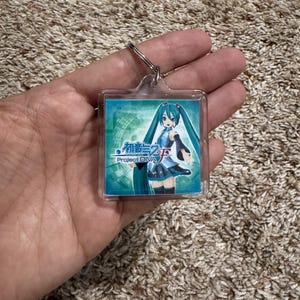 May include: A square acrylic keychain featuring a graphic of the character Hatsune Miku with long teal pigtails, wearing a blue and black outfit. The image includes the text Hatsune Miku Project DIVA F in blue and white lettering on a teal background.
