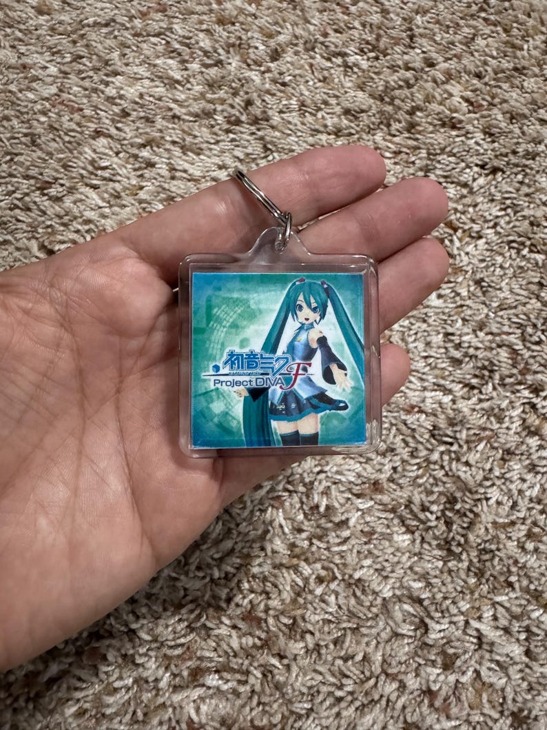 May include: A square acrylic keychain featuring a graphic of the character Hatsune Miku with long teal pigtails, wearing a blue and black outfit. The image includes the text Hatsune Miku Project DIVA F in blue and white lettering on a teal background.