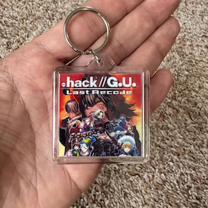 Pode incluir: Um chaveiro quadrado transparente com o texto "hack //G.U. Last Recode" em vermelho e branco. O chaveiro tem um anel de metal e um gr&aacute;fico colorido de personagens do jogo.