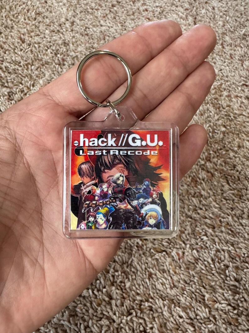 Pode incluir: Um chaveiro quadrado transparente com o texto "hack //G.U. Last Recode" em vermelho e branco. O chaveiro tem um anel de metal e um gr&aacute;fico colorido de personagens do jogo.