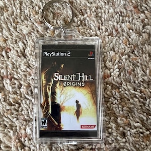 May include: A clear acrylic keychain featuring a PlayStation 2 game case for "Silent Hill: Origins." The game case art depicts a figure in a dark forest. The keychain has a silver metal ring.
