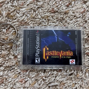 May include: A Castlevania: Symphony of the Night PlayStation game keychain. The keychain features the game's cover art in a clear plastic case with a silver key ring.
