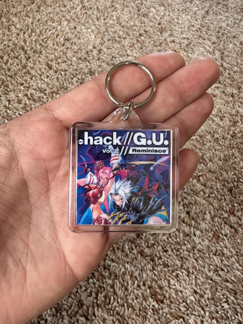 Pode incluir: Um chaveiro de acr&iacute;lico transparente com o texto "hack//G.U. Vol. 2 Reminisce" e uma ilustra&ccedil;&atilde;o colorida em estilo anime. Possui um anel de metal prateado.