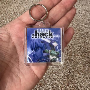 Pode incluir: Um chaveiro quadrado transparente com o texto "hack OUTBREAK Part 3" e caracteres japoneses. A imagem no chaveiro mostra uma ilustra&ccedil;&atilde;o em estilo anime em tons de azul de um personagem com apar&ecirc;ncia de coelho.