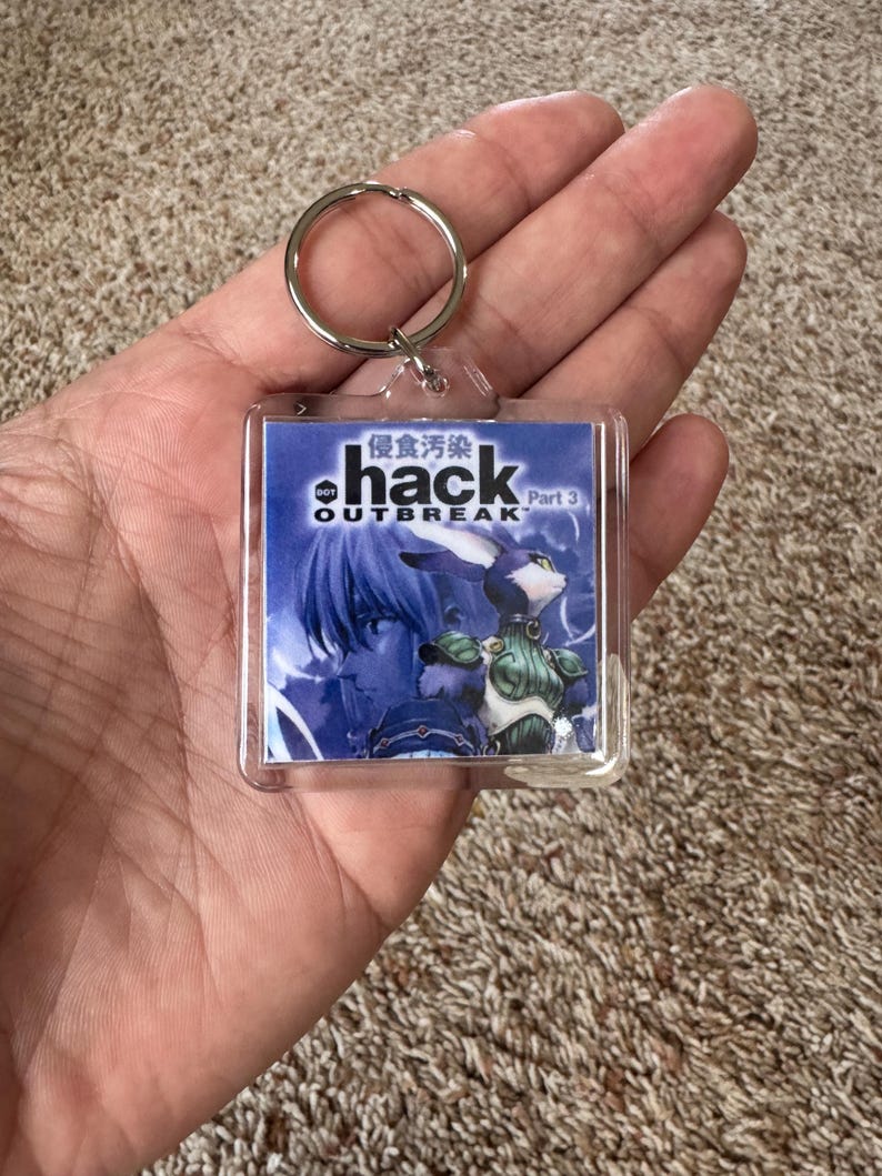 Pode incluir: Um chaveiro quadrado transparente com o texto "hack OUTBREAK Part 3" e caracteres japoneses. A imagem no chaveiro mostra uma ilustra&ccedil;&atilde;o em estilo anime em tons de azul de um personagem com apar&ecirc;ncia de coelho.