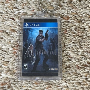 May include: A clear acrylic keychain featuring the Resident Evil 4 PS4 game cover art. The image shows a man holding a gun, with the game title and the Capcom logo. The keychain has a silver metal ring.