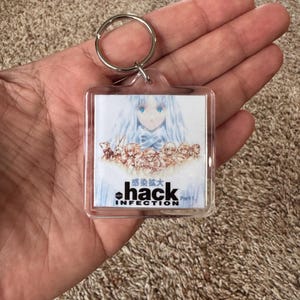 Pode incluir: Chaveiro quadrado transparente com arte em estilo anime. A imagem mostra uma personagem com cabelo comprido azul e um la&ccedil;o azul. O texto "hack INFECTION" &eacute; impresso abaixo, juntamente com caracteres japoneses.