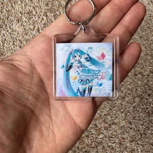 May include: A square clear acrylic keychain featuring an illustration of the character Hatsune Miku with teal hair. The image includes the text Hatsune Miku Project DIVA f 2nd, musical notes, and geometric shapes against a light blue and purple background.