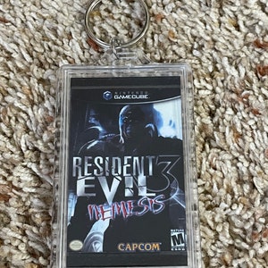May include: Clear acrylic keychain featuring the Resident Evil 3: Nemesis video game cover art. The keychain includes a silver metal ring for attaching to keys or bags. The game cover art features a dark background with the game title and a menacing character.