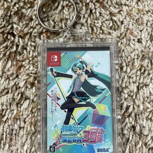 May include: A clear acrylic keychain featuring a colorful graphic of a character from the game Hatsune Miku: Project DIVA Mega 39's. The keychain has a silver metal ring and a rectangular design with Japanese text and the Nintendo Switch logo.