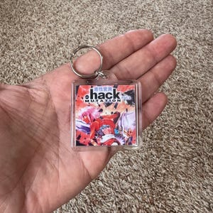 Pode incluir: Um chaveiro quadrado com um anel de metal. O chaveiro tem um gr&aacute;fico colorido com a palavra "hack" em uma fonte estilizada e texto japon&ecirc;s. O gr&aacute;fico apresenta personagens em estilo anime em vermelho, rosa e azul.