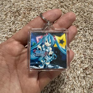 May include: A square clear acrylic keychain featuring a printed graphic of the character Hatsune Miku with long teal twin tails, wearing a metallic blue outfit. The background includes a yellow star and the text Hatsune Miku Project DIVA F.