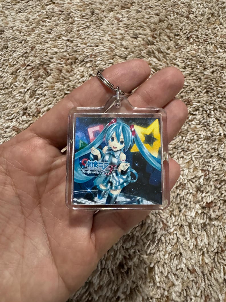 May include: A square clear acrylic keychain featuring a printed graphic of the character Hatsune Miku with long teal twin tails, wearing a metallic blue outfit. The background includes a yellow star and the text Hatsune Miku Project DIVA F.