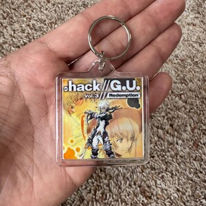 Pode incluir: Um chaveiro quadrado de acr&iacute;lico transparente com uma argola prateada, apresentando arte de "hack//G.U. Vol. 3 Redemption". O chaveiro exibe uma ilustra&ccedil;&atilde;o colorida em estilo anime com o t&iacute;tulo e o n&uacute;mero do volume do jogo.