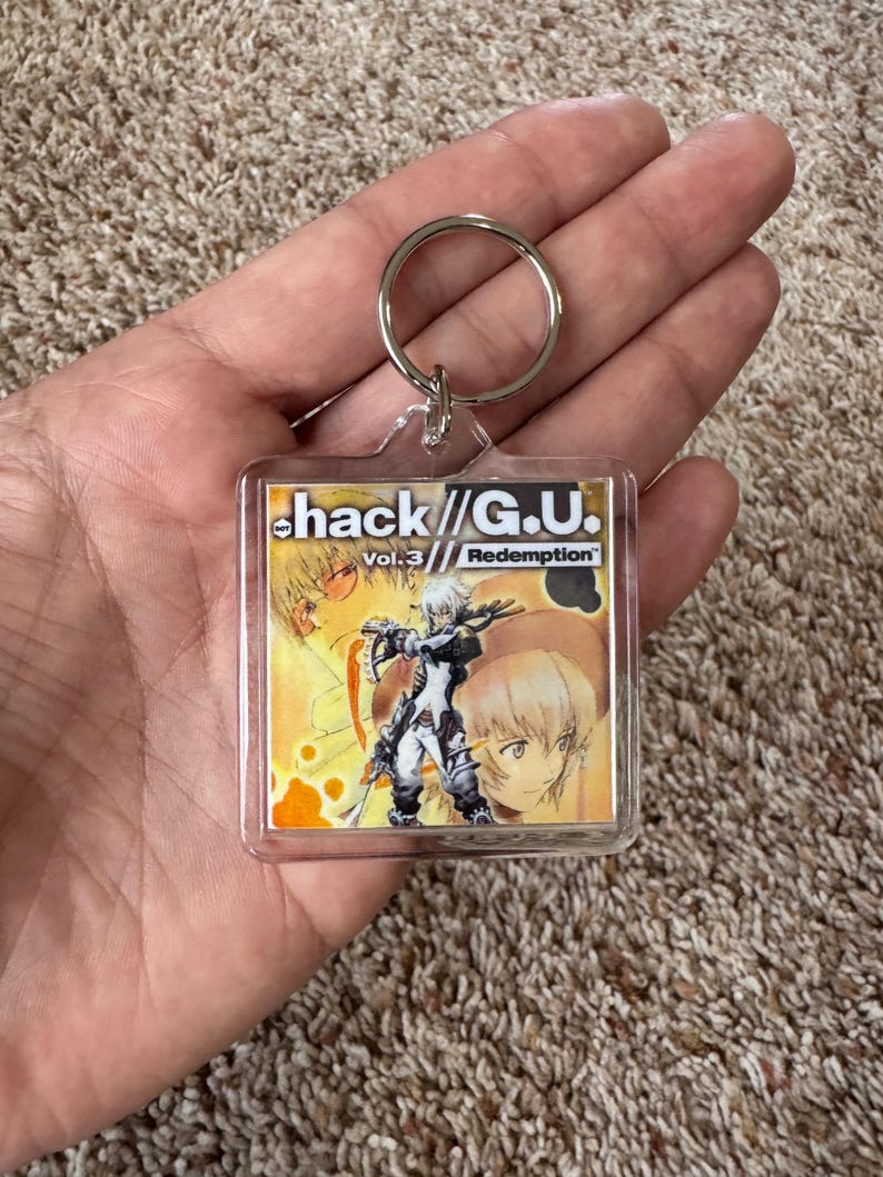 Pode incluir: Um chaveiro quadrado de acr&iacute;lico transparente com uma argola prateada, apresentando arte de "hack//G.U. Vol. 3 Redemption". O chaveiro exibe uma ilustra&ccedil;&atilde;o colorida em estilo anime com o t&iacute;tulo e o n&uacute;mero do volume do jogo.