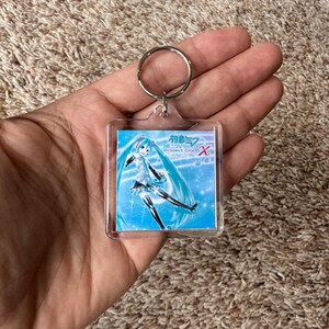 May include: A square clear acrylic keychain featuring a printed graphic of the character Hatsune Miku with long teal hair and a blue outfit. The image includes the text Hatsune Miku Project DIVA X in white and red lettering against a blue background.