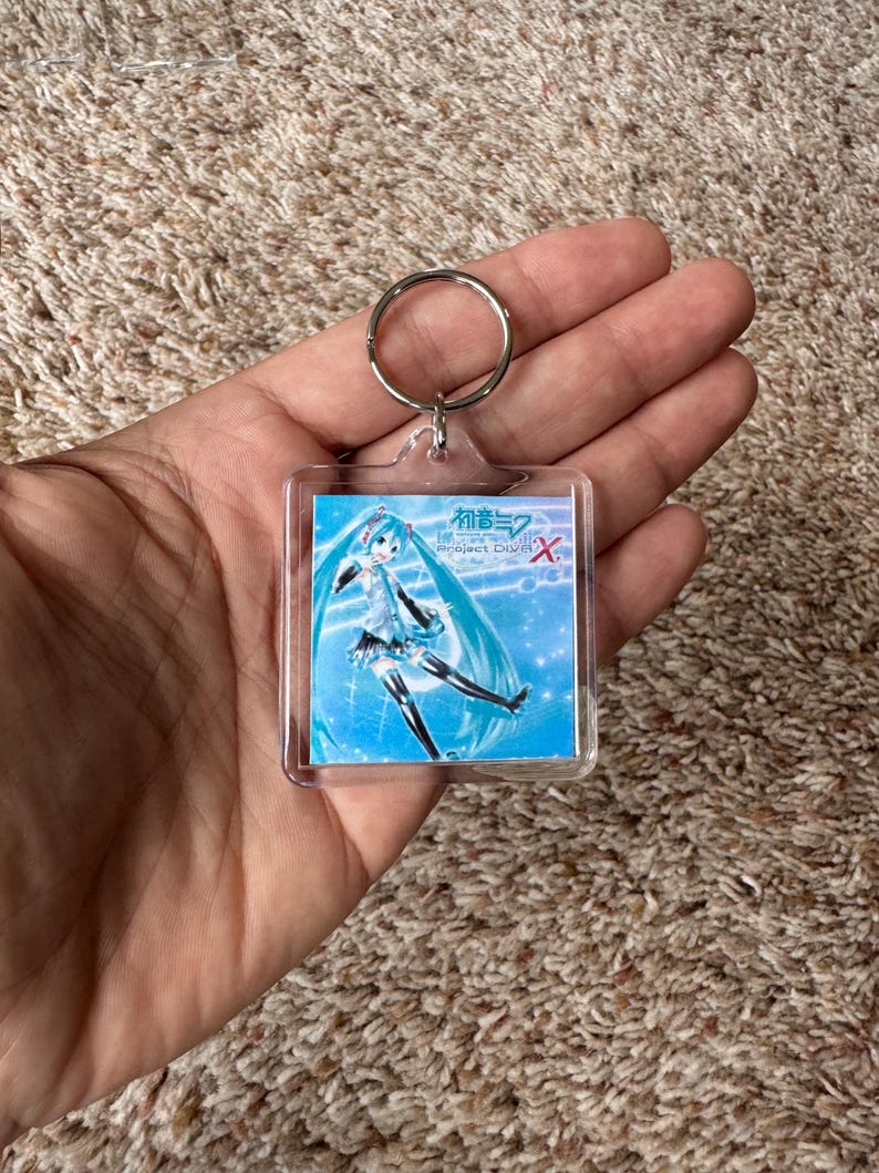 May include: A square clear acrylic keychain featuring a printed graphic of the character Hatsune Miku with long teal hair and a blue outfit. The image includes the text Hatsune Miku Project DIVA X in white and red lettering against a blue background.