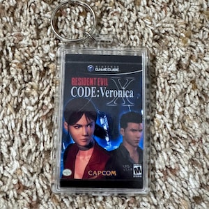 May include: A clear acrylic keychain featuring the Resident Evil Code: Veronica video game cover art. The artwork shows two characters in a dark setting, with the Capcom logo at the bottom. The keychain has a silver metal ring.