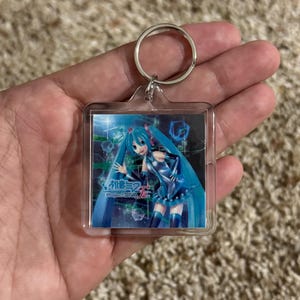 May include: A square clear acrylic keychain featuring a printed graphic of the character Hatsune Miku with long teal hair and a blue outfit. The image includes the text Hatsune Miku Project DIVA F 2nd set against a dark digital background.