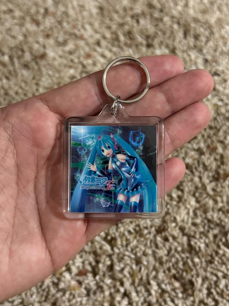 May include: A square clear acrylic keychain featuring a printed graphic of the character Hatsune Miku with long teal hair and a blue outfit. The image includes the text Hatsune Miku Project DIVA F 2nd set against a dark digital background.