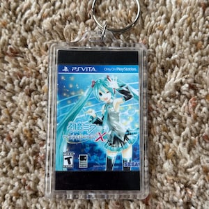 May include: Clear acrylic keychain featuring a PS Vita game cartridge design. The game cover art showcases a teal-haired anime character with the text "Project DIVA X" and the SEGA logo. Includes a silver keyring.
