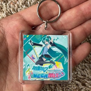 May include: A square acrylic keychain featuring a colorful illustration of the character Hatsune Miku with teal hair, wearing a blue and black outfit. The design includes the text Hatsune Miku Project DIVA MEGA MIX and musical note symbols.