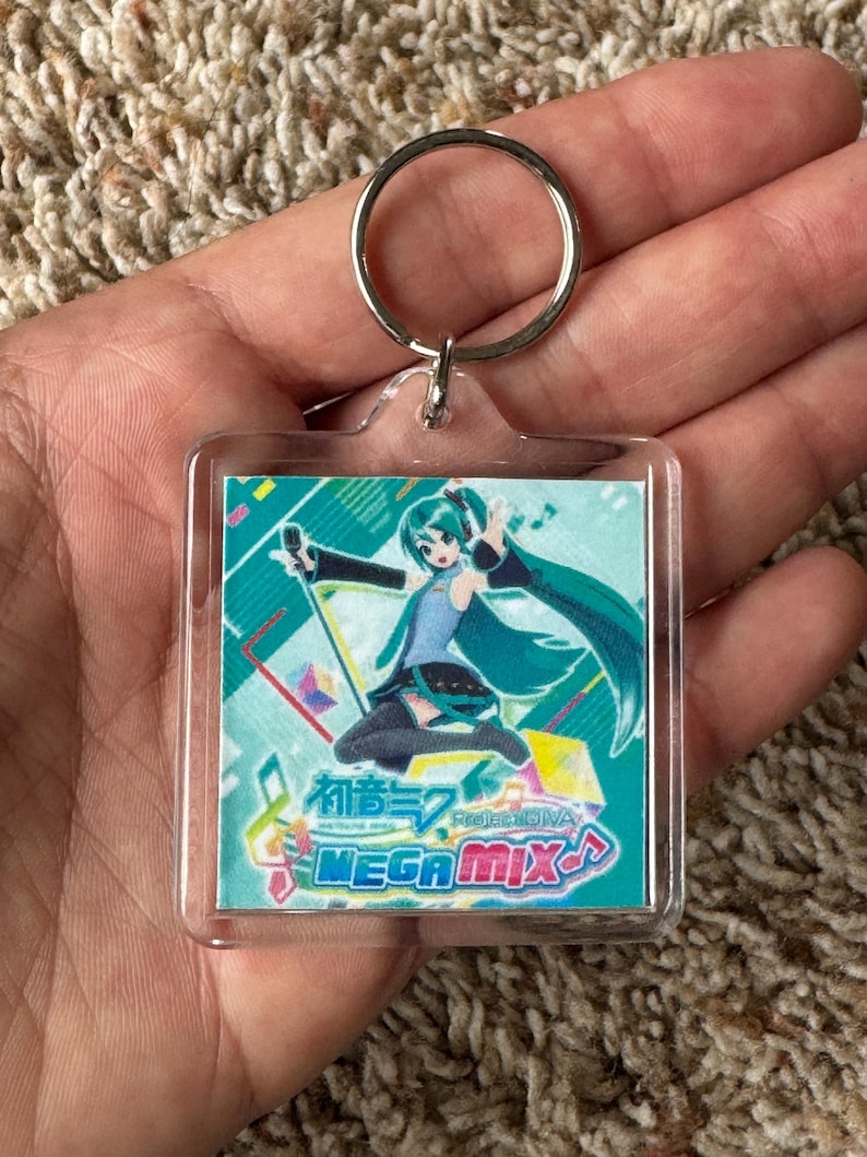 May include: A square acrylic keychain featuring a colorful illustration of the character Hatsune Miku with teal hair, wearing a blue and black outfit. The design includes the text Hatsune Miku Project DIVA MEGA MIX and musical note symbols.